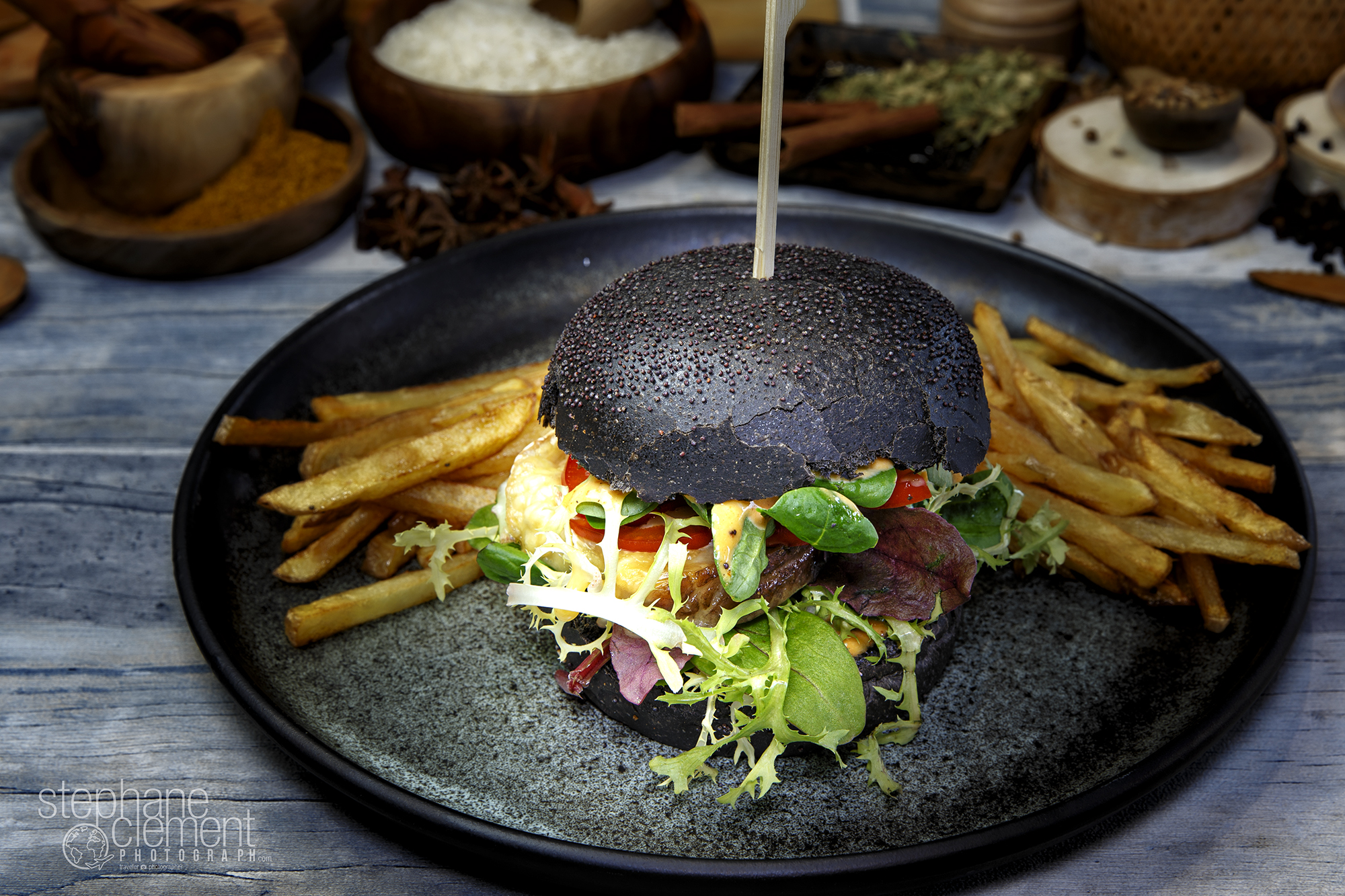 Black Burger with salad, cheese, tomato and beef, accompanied by French fries on a white plate, served on a design plate. Decoration in background with cooking tools.