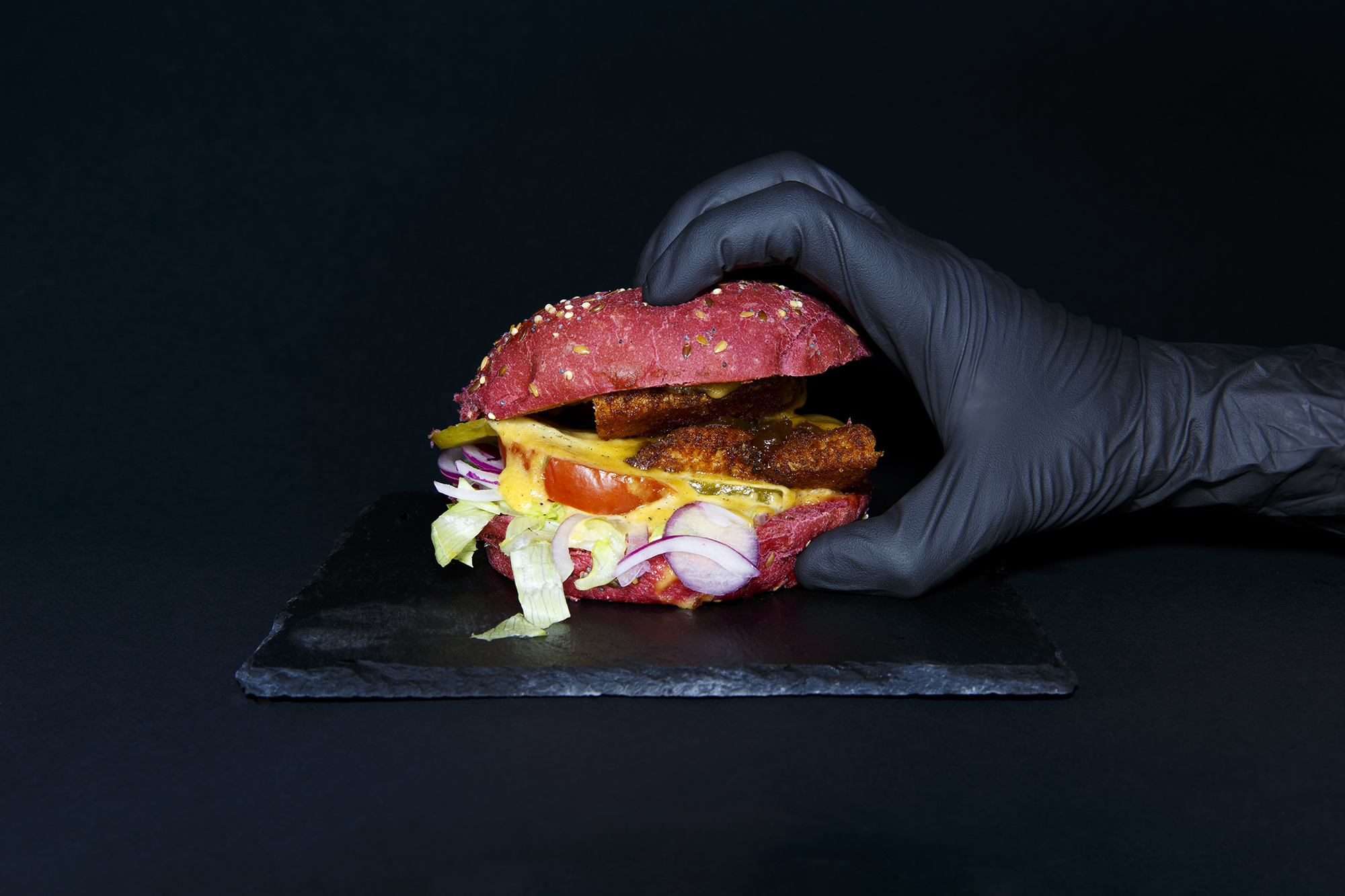 Pink burger with soft bread, breaded chicken, homemade cheddar sauce, candied onions, salad, tomatoes. Served in a slate on a black background. Hand of a woman holding a fork in the plate.