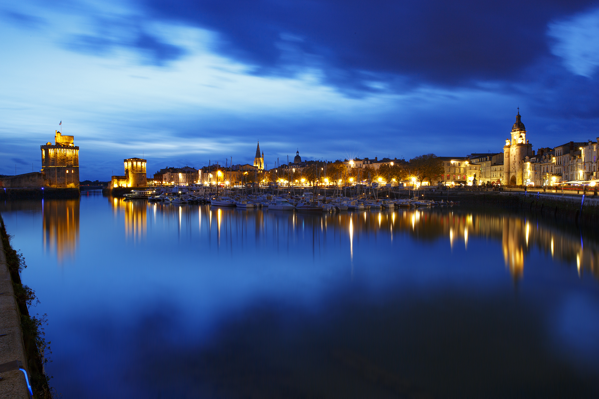 The city of La Rochelle in France, during the blue hour, view of the port with the towers of the entrance to the marina. Buildings, restaurants and hotels with illuminated facades. November 2022.