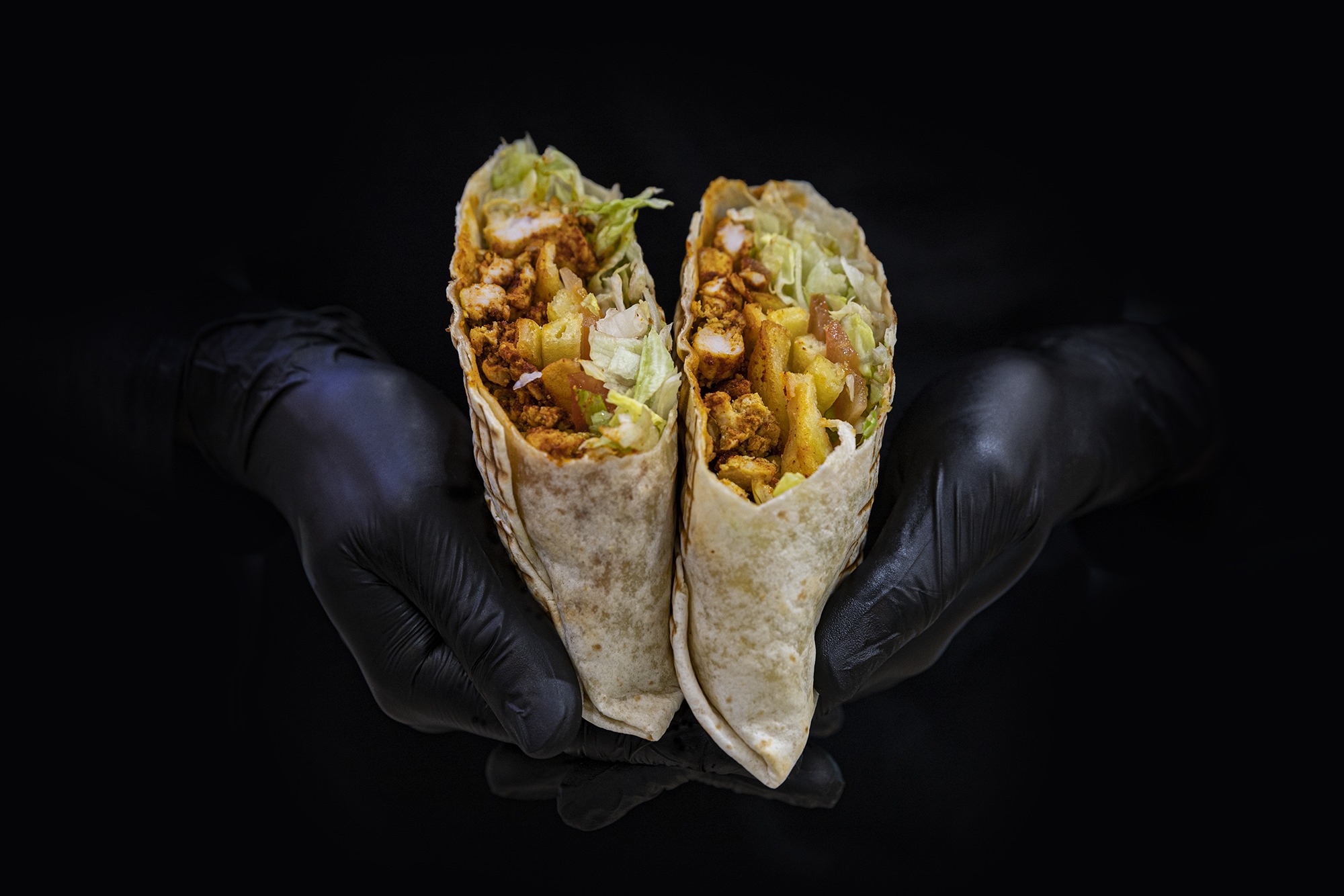 Main de cuisinier avec gants en latex noir qui tient un tacos coupé en deux.