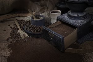 Scène de café avec du café en grain, des spatules en bois et un vieux moulin à café manuel en fonte. Mise en scène sur une table en bois avec du café moulu, des grains de café et un vieux sac en toile en arrière plan. 2 tasses à café sont dispo